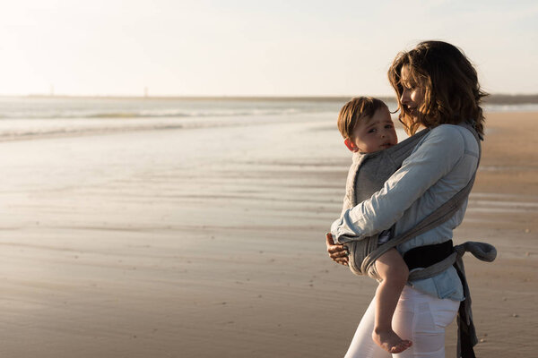 Мать с эргобэби несет малыша на пляже
