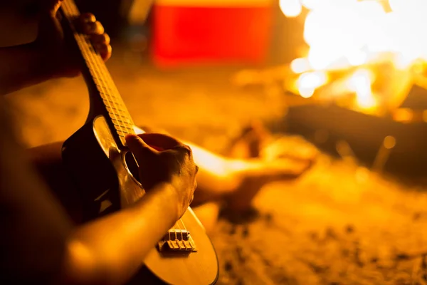 Man tokkelen een ukulele/gitaar in het bos naast een open vuur — Stockfoto