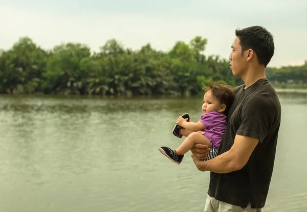 Asiático pai está levando seu filho para um passeio ao longo do lago carregando a criança em seus braços — Fotografia de Stock