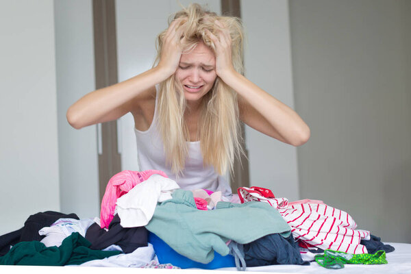 A depressed exhasted woman sitting on her bed with a pile of laundry, not happy to do housework at home.
