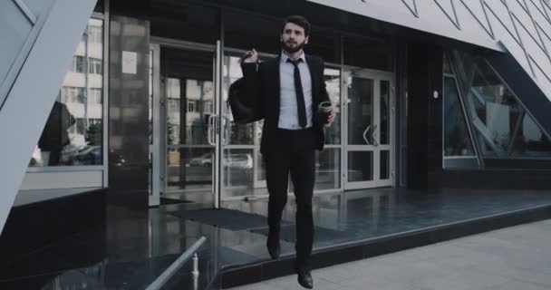 Молодой бизнесмен закончил свой рабочий день, выходя из современного здания с кофе и портфелем — стоковое видео