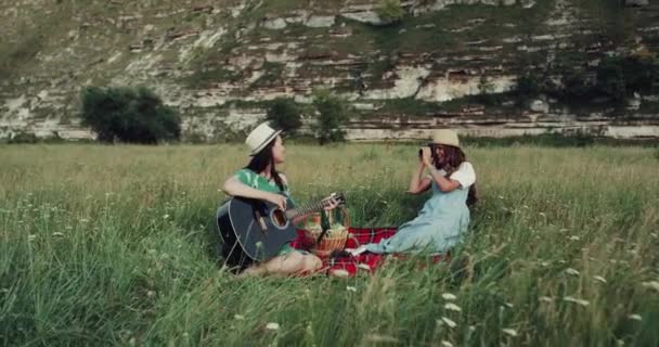 Dos chicas tienen picnic en el lugar increíble con montañas, están cantando en la guitarra y tomando fotos increíbles. mociones lentas — Vídeos de Stock