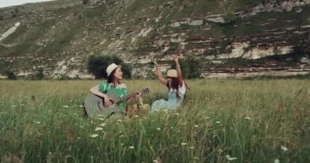 Två flickor dans och sång på gitarr, mitt i naturen fastställande på filten — Stockvideo