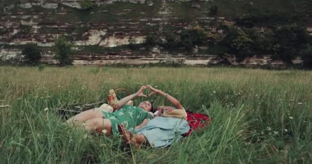 Hora del picnic con dos chicas en medio de la montaña acostadas sobre la manta y haciendo corazón con las manos. 4k — Vídeos de Stock