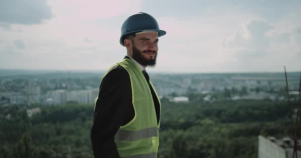 Счастливый и уверенный в себе архитектор с защитным шлемом на стройке, улыбаясь в камеру. 4k. замедленное движение — стоковое видео