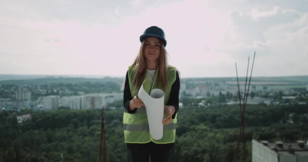 Unga vackra kvinnliga arkitekt med en skyddshjälm stå på toppen av konstruktion och ler mot kameran. — Stockvideo