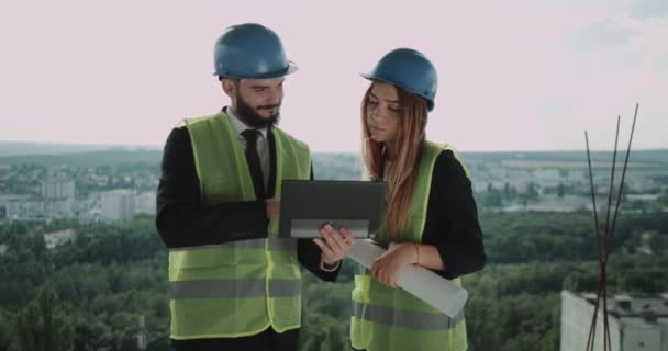 Δύο νεαροί μηχανικοί άνδρα και γυναίκα έχουν μια συζήτηση στην κορυφή του εργοταξίου, χρησιμοποιώντας ένα tablet αναλύοντας το σχέδιο του κτιρίου. — Αρχείο Βίντεο
