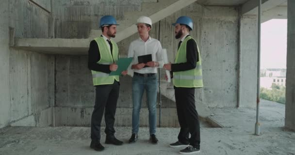 Drie jonge deskundige ingenieurs analyseren van de kaart van nieuw gebouw met behulp van een tablet, het dragen van een veiligheidshelm en hoge zichtbaarheid vesten — Stockvideo