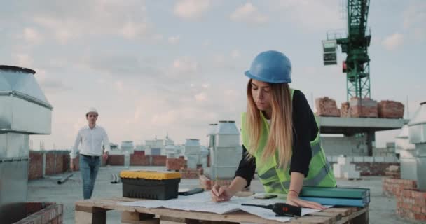 Jóvenes ingenieros analizan plan de construcción en la azotea de la construcción, los trabajadores de la construcción de fondo discutiendo juntos . — Vídeos de Stock