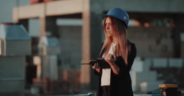 在屋顶上建设一个安全头盔的年轻女子, 用无线电说话, 并通过平板电脑查看. — 图库视频影像