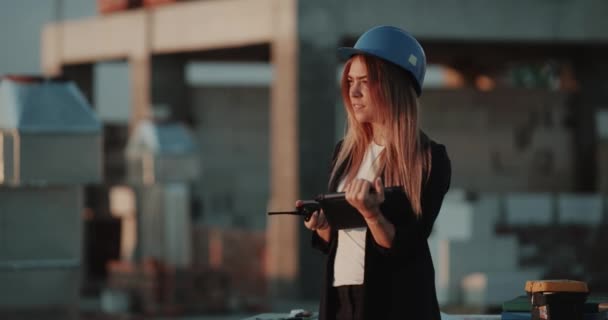 En la azotea de la construcción de la mujer joven con un casco de seguridad, hablando con una radio y mirando a través de la tableta . — Vídeo de stock