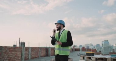 Çatı katında inşaat site işadamı bir kask ve bir siyah radyosunun kullanımının yüksek görünürlük yelek konuşma giyiyor.