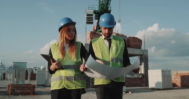 Twee partners uit bouwplaats lopen rond het gebouw op het dak en het plan van een nieuwe appartementen, op achtergrond hoge kraan te bespreken. 4k — Stockvideo