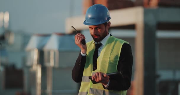 Jongeman supervisor bij bouwplaats spreken van radio en het analyseren van het plan van de bouw van tablet. — Stockvideo