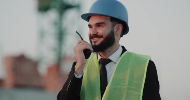 Νέος μηχανικός στο εργοτάξιο φορώντας κράνος ασφαλείας και μιλώντας με εργάτες του χρησιμοποιώντας ένα ραδιόφωνο — Αρχείο Βίντεο