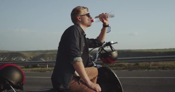 Uma menina de cabelos ruivos latina está sentada em uma moto e está bebendo uma garrafa de água na estrada — Vídeo de Stock