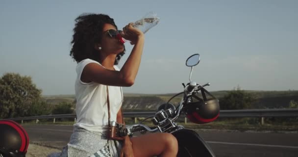 Una chica latina de pelo rizado tiene sed bebiendo su agua y está sentada en su bicicleta junto a la carretera. — Vídeos de Stock