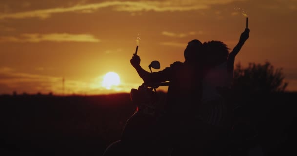 Al atardecer, la joven pareja se divierte detrás de su motocicleta retro negra, sosteniendo los bengalas y disfrutando del tiempo juntos. 4k — Vídeo de stock