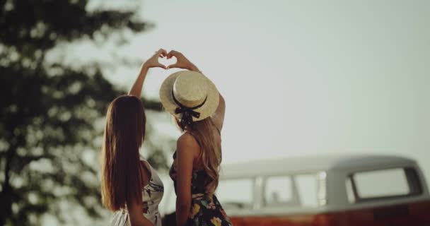 Joven dos mujeres de 90 s en medio de la naturaleza con su autobús retro, haciendo un corazón latiendo en el aire con las manos . — Vídeo de stock