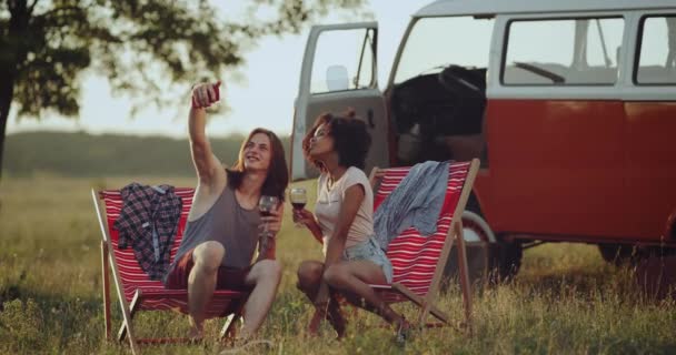 Beautiful couple, have a picnic time in the amazing place under the beautiful sky, sitting on wooden chairs drinking wine, taking selfies, background amazing retro van . — стоковое видео