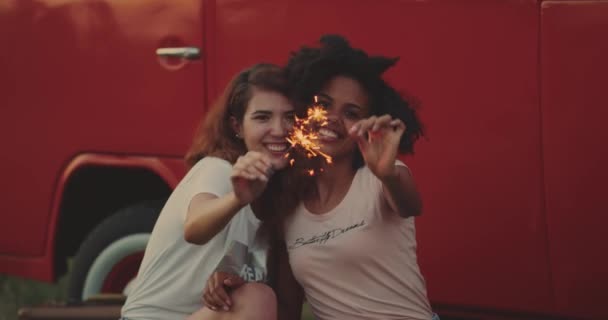 Uma jovem africana e sua amiga, comprada com cabelo encaracolado sorrindo brincando com um espumante no piquenique sentado na grama ao lado de uma van retro. 4k . — Vídeo de Stock