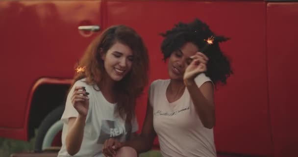 Bela jovem africana e sua amiga bonita se divertem juntos na natureza brincando com espumantes sentados ao lado de uma van vintage . — Vídeo de Stock