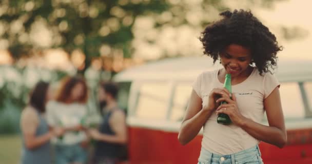 Mujer joven africana con el pelo rizado de cerca en el picnic muy carismático y hermoso mirando directamente a la cámara sonriendo, y sosteniendo una botella verde . — Vídeo de stock