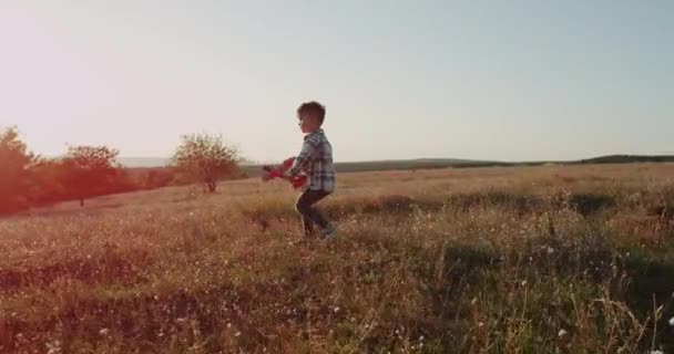 Niño sonriente jugando con un avión, en medio del campo, saltando feliz, hermoso atardecer . — Vídeos de Stock