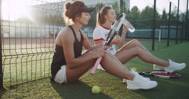 En la cancha de tenis por la noche dos chicas relajándose después del juego bebiendo una batalla de agua . — Vídeo de stock