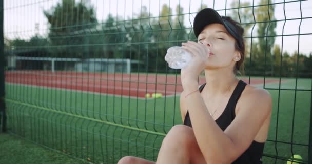 Senhora sorridente carismática bebendo uma garrafa de água sentada na grama na quadra de tênis, antes de iniciar o jogo de tênis . — Vídeo de Stock