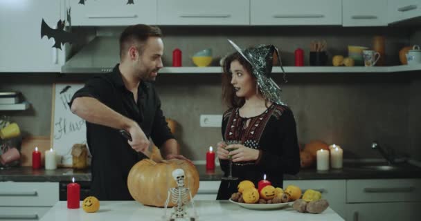 若いカップルの素晴らしい時間を過ごす大きなハロウィンかぼちゃをカットしようとしてキッチン食卓でハロウィン パーティーの準備を笑顔女性カクテルのグリーン ガラスを既に保持しているマスクは — ストック動画