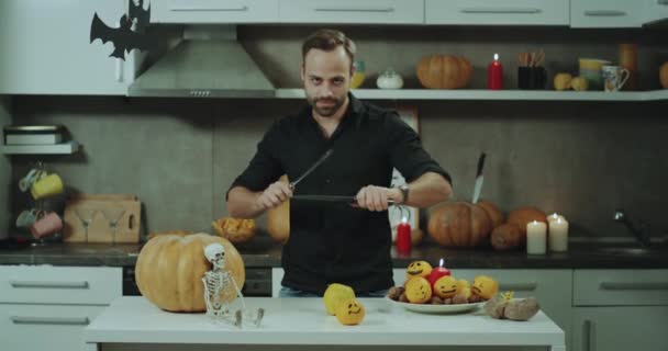 Serios hombre con una barba jugando con cuchillos en su mesa de la cocina se preparan para cortar una calabaza grande de Halloween. — Vídeo de stock