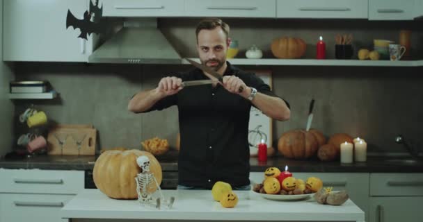 Sorridente giovane uomo in una maglietta nera che gioca con coltelli alla sua cucina prima di una festa di Halloween, vuole tagliare una zucca. 4k — Video Stock