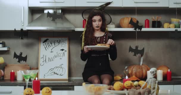 Молодая ведьма в маске сидит на кухонном столе и держит в руках тарелку с закусками, играющую перед камерой . — стоковое видео