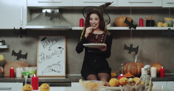 Uma bruxa com lábios pretos comendo soo engraçado o lanche na festa de Halloween, ela está sentada na mesa da cozinha e segurando o prato, tem um grande sorriso branco. Tiro na Red Epic. movimentos lentos . — Vídeo de Stock