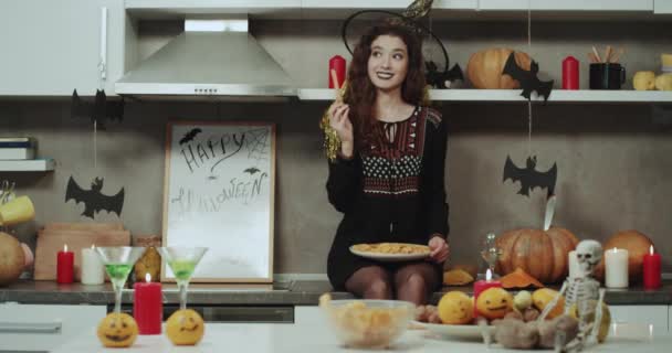 Eine lustige und schöne maskierte Hexe in der Halloween-Nacht hat gute Laune in der Küche und hält einen Teller mit Snacks in die Kamera. — Stockvideo