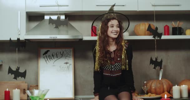 Genç Bayan mutfak masasının üstünde duran ve için oynayan bir cadı maskeli çok tabir yüzü kameranın Cadılar Bayramı gecesi. 4k — Stok video