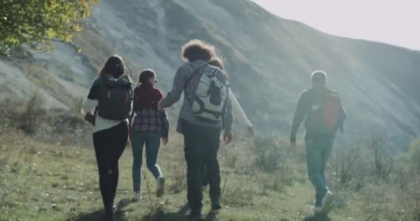 Um grupo amigável de jovens tem um tempo de viagem caminhando através do campo para a montanha, eles têm um grande sacos de viagem em suas costas . — Vídeo de Stock