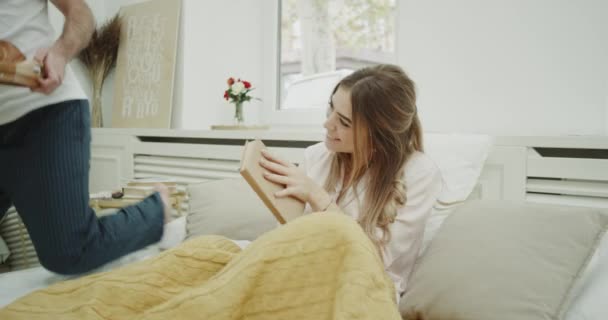 舒适的卧室在早晨一个漂亮的女士正在看书, 她的男人在床上吃早餐感到惊讶. — 图库视频影像