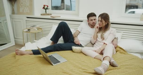 Pareja joven en apartamento moderno divertirse juntos en el dormitorio comer palomitas de maíz en la cama y ver películas en el ordenador portátil . — Vídeos de Stock