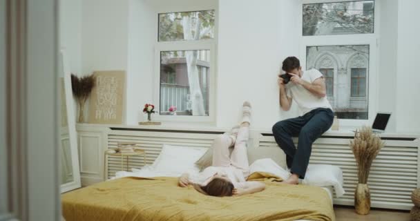 Jonge kerel maken foto's voor zijn vriendin in hun gezellige slaapkamer, ze is vaststelling van op het bed en het dragen van een schattig roze pyjama's. 4k — Stockvideo