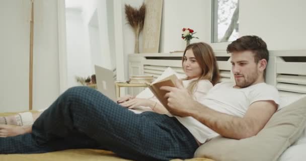 Em um quarto moderno carismático sorrindo jovem casal ter um tempo frio na cama em pijama livro de leitura e olhando através do caderno . — Vídeo de Stock