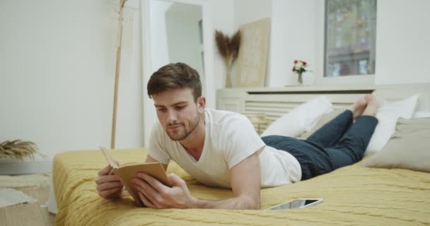 Junger Mann im Schlafanzug liest abends im Bett ein Buch. — Stockvideo