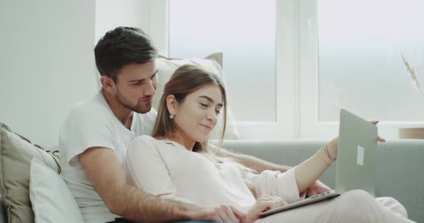 Retrato de pareja joven pedir algo usando un cuaderno sentado en el sofá y ambos en pijama — Vídeos de Stock