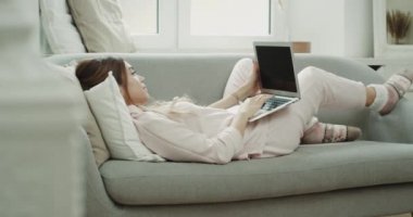 Bayan kanepede rahat tarzı bir pijama giymiş bir dizüstü bilgisayar üzerinde çalışan.