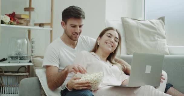 Week-end pour un jeune couple passer un bon moment ensemble dans le salon assis sur le canapé manger du pop-corn et regarder un film de portable . — Video