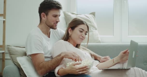 Geweldige tijd doorbrengen paar samen thuis kijken naar film van laptop en eten popcorn, in de moderne woonkamer zowel in pyjama. — Stockvideo