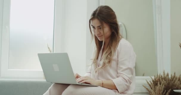 In een gezellige woonkamer met een mooie jonge dame die werken op een laptop zitten in de Bank en het dragen van een pyjama. 4k. geschoten op het rode epos — Stockvideo