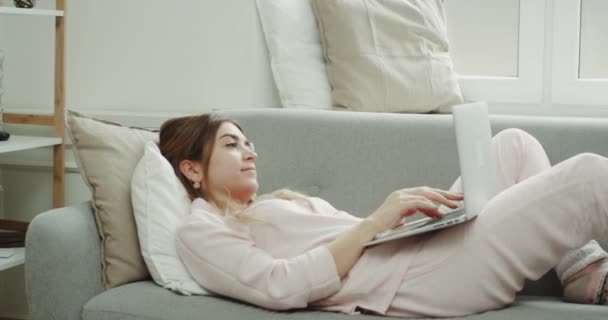 Diseño moderno en la sala de estar mujer joven tocando en el portátil que está sentado en el sofá en pijama . — Vídeo de stock
