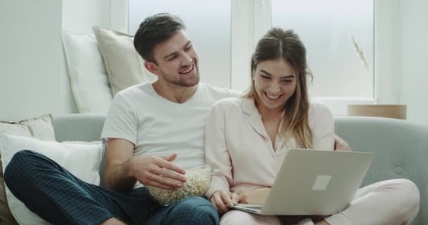 Preciosa pareja feliz y sonriente comer palomitas de maíz y ver algo en el portátil, sentado en el sofá de estilo acogedor . — Vídeos de Stock